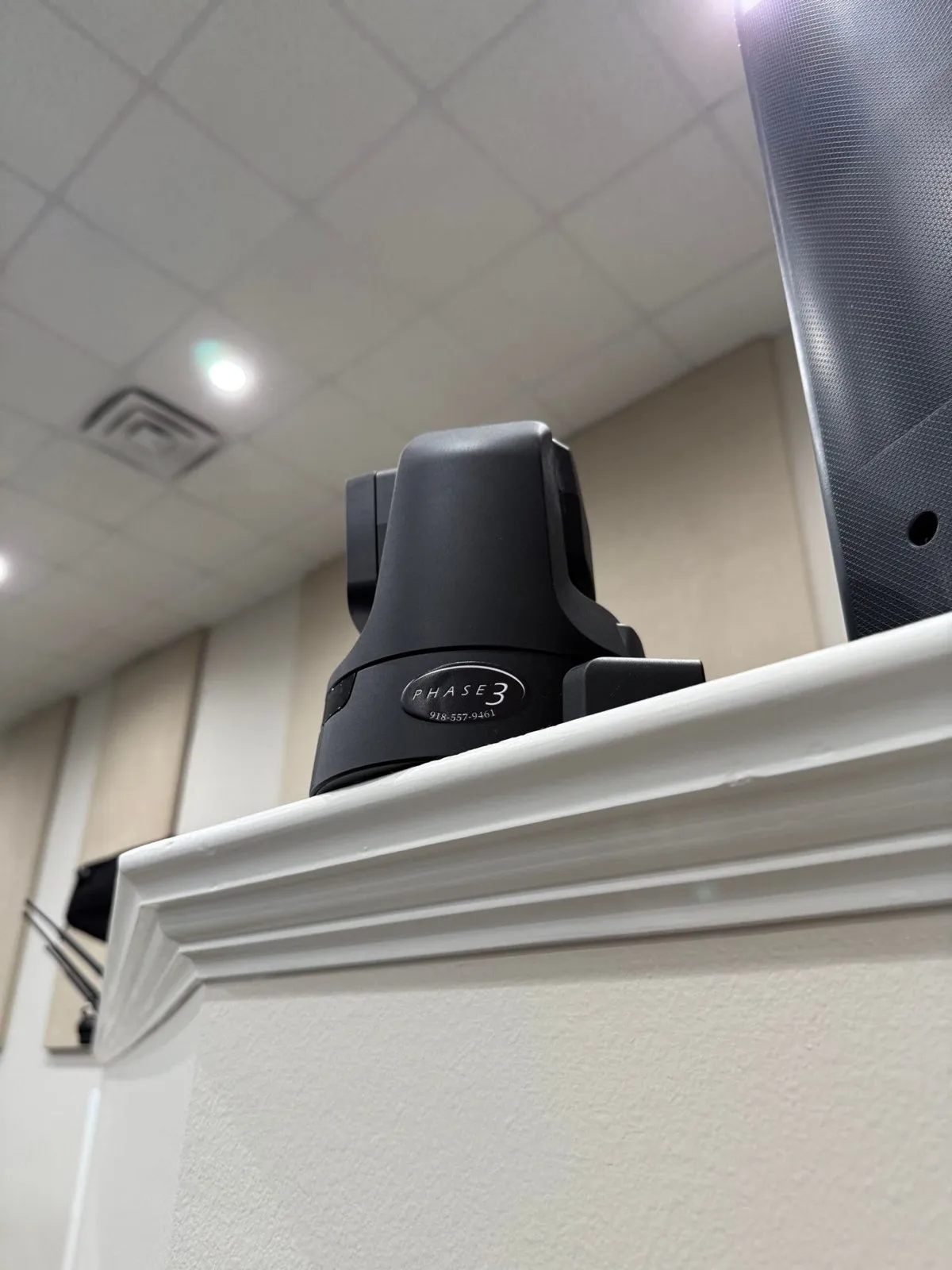PTZ video camera with Phase 3 Live branding installed on a ledge next to a loudspeaker in a worship venue.
