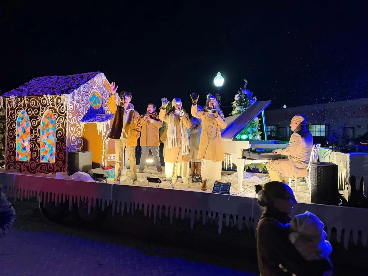 Performers singing on a gingerbread-house Christmas parade float with live sound support at a nighttime holiday event.