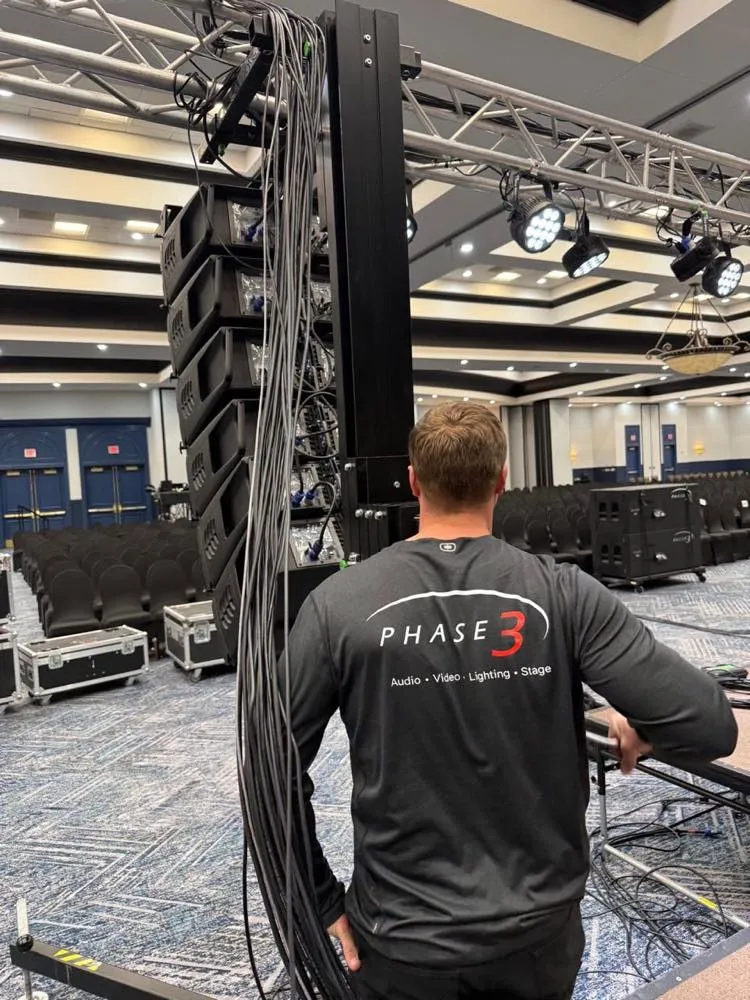 Phase 3 Live crew member managing cabling at the base of a line array tower during a ballroom event setup.