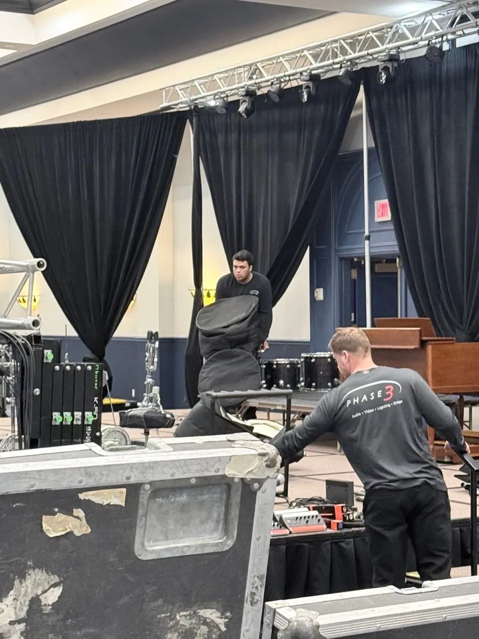 Phase 3 Live crew packing instrument cases on stage in front of black pipe and drape