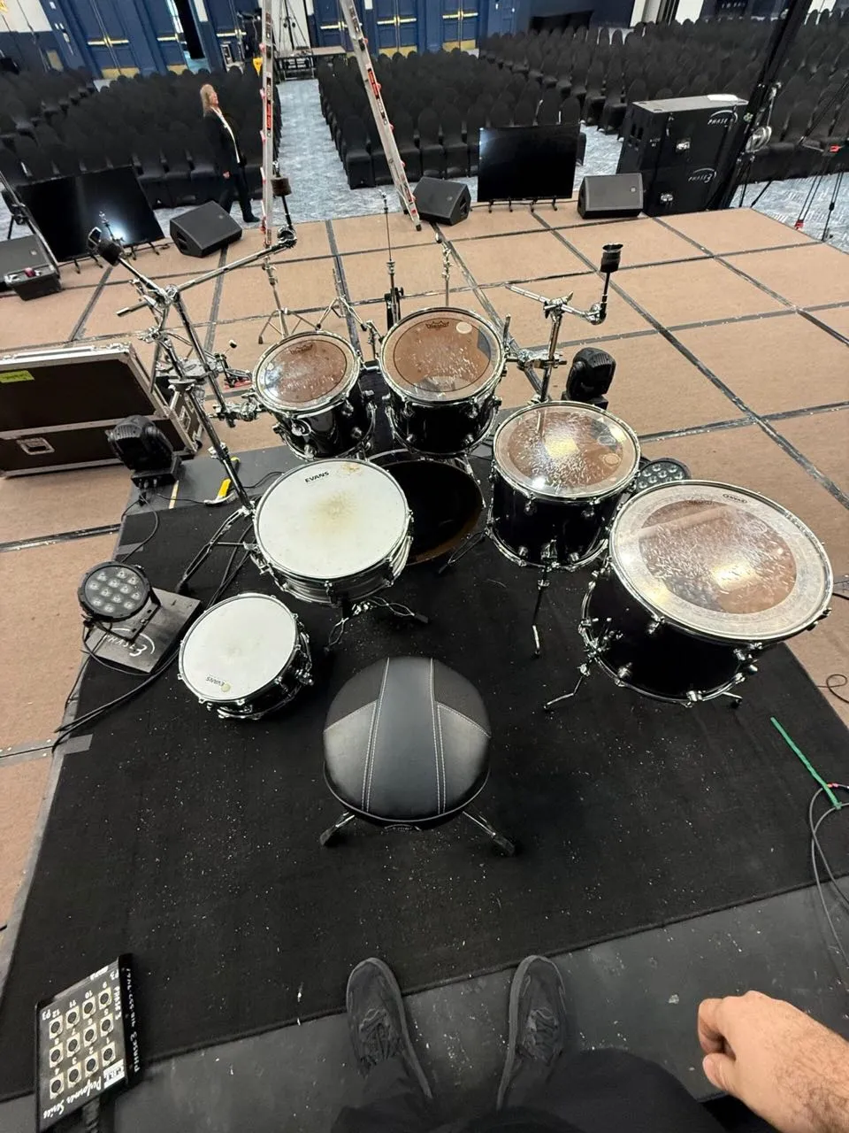 View from behind a drum kit on a stage riser set by Phase 3 Live facing an empty ballroom.