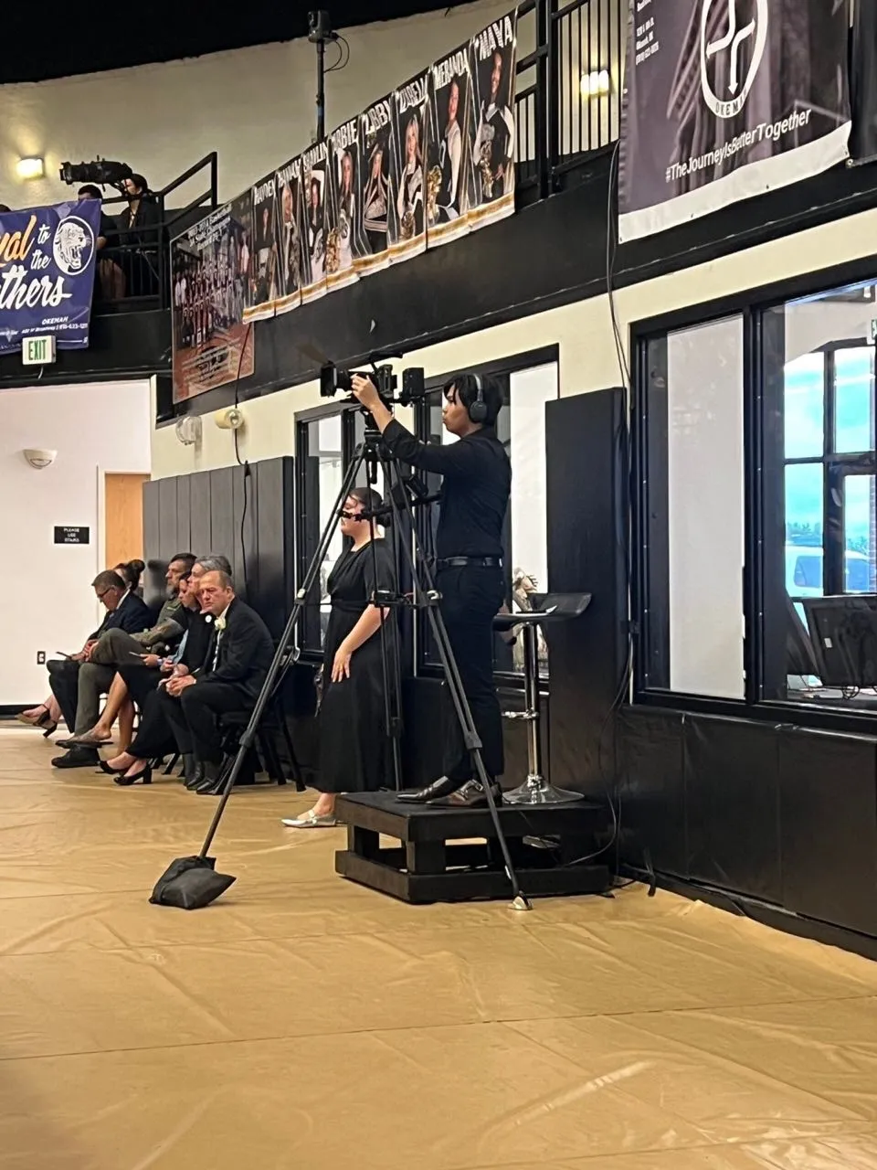 Camera operator filming an indoor ceremony from a riser as seated guests look on