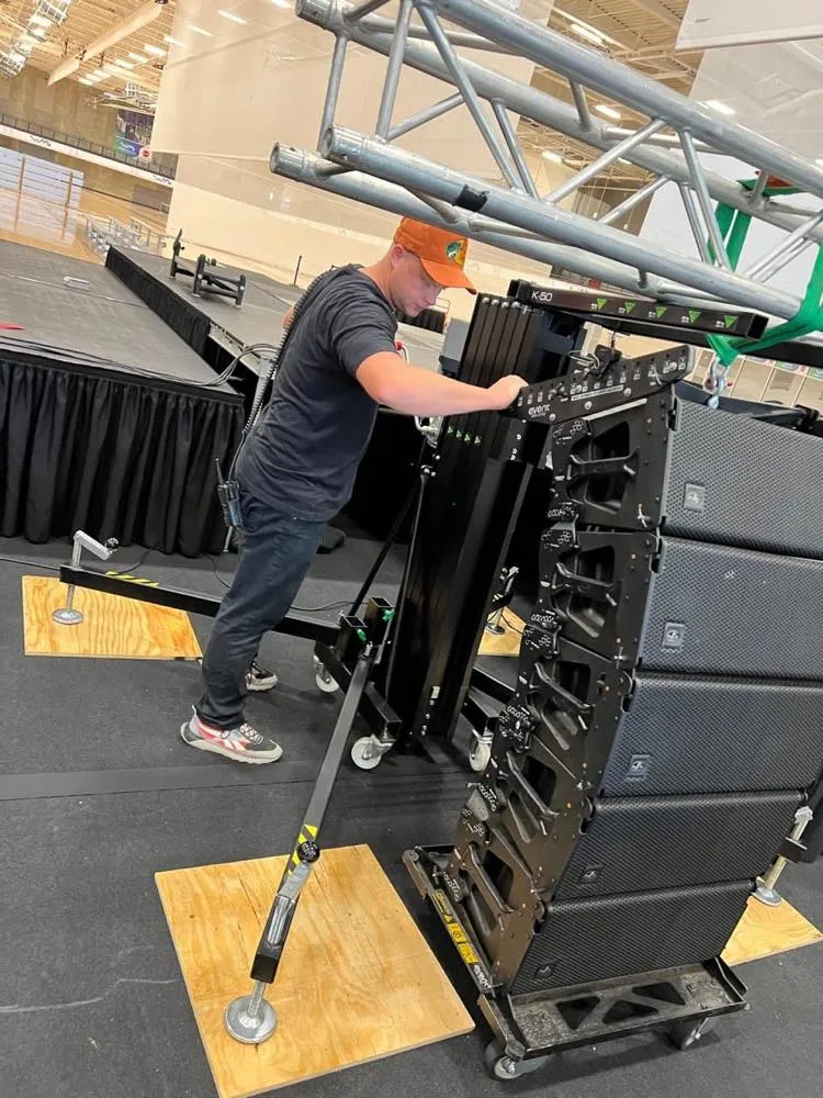 Phase 3 Live technician assembling a line array on a crank-up lift tower beside an event stage