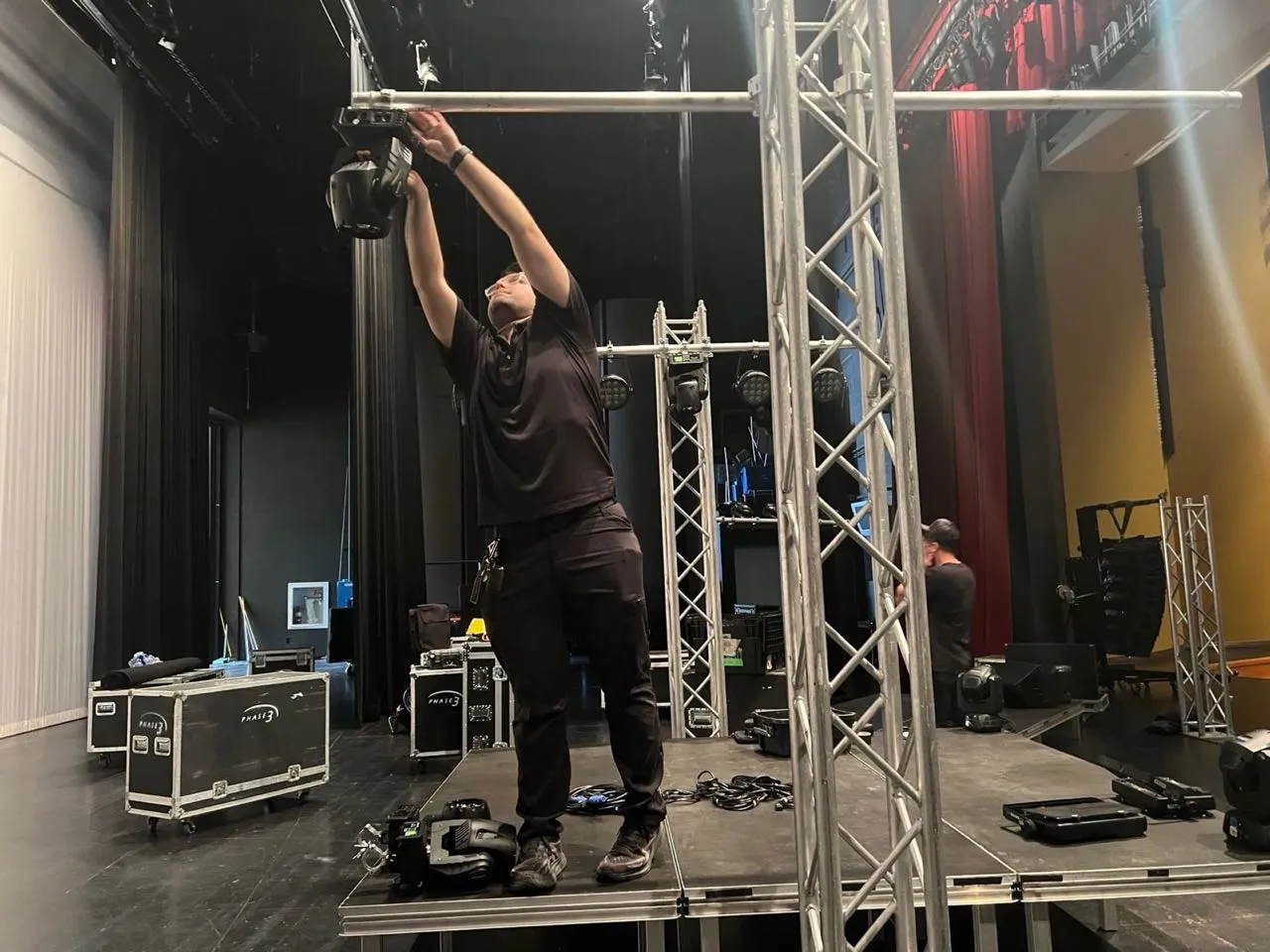 Phase 3 Live crew member installing a moving-head lighting fixture on truss in a theater