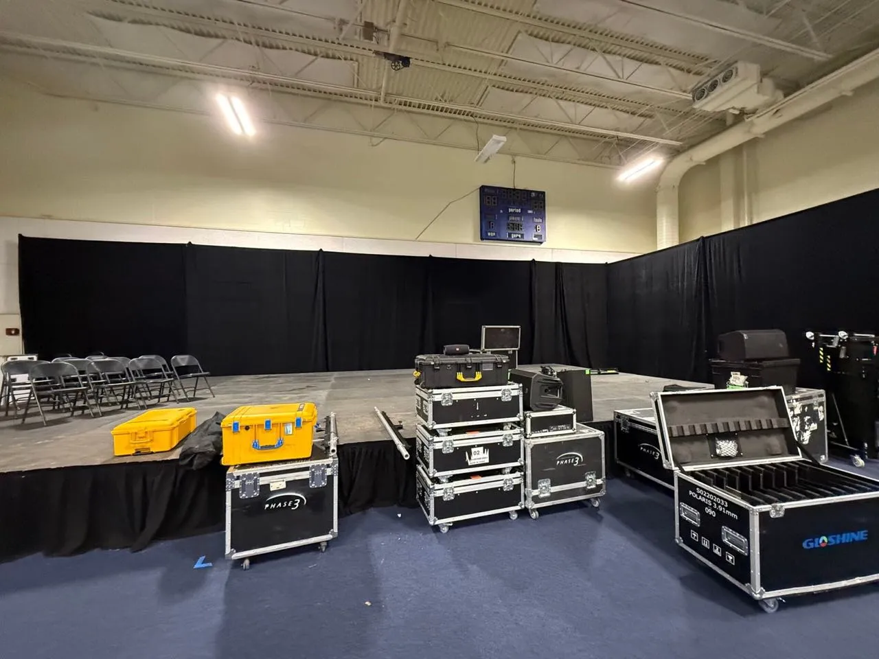Gymnasium stage framed with black pipe and drape, with Phase 3 Live road cases and LED panel cases in front