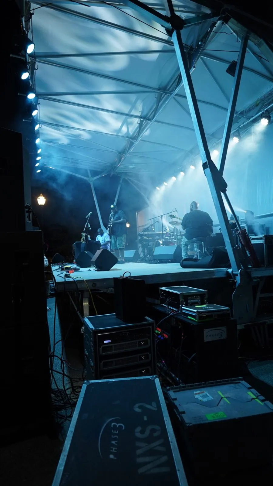Phase 3 Live road cases and amp racks behind an outdoor stage during a live band performance