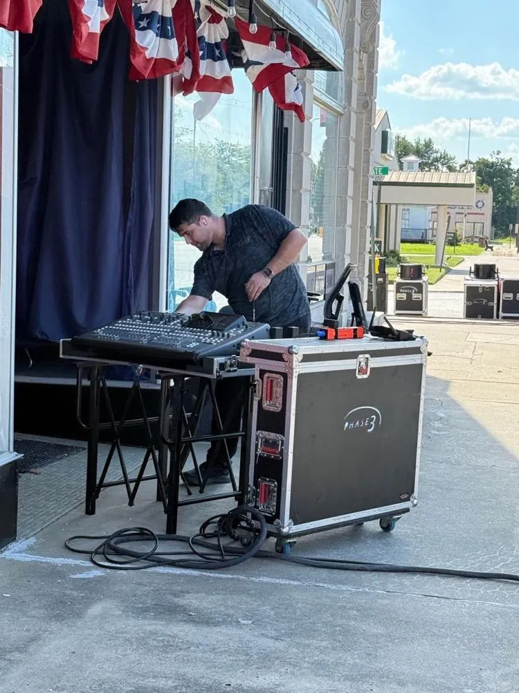 Phase 3 Live audio engineer setting up a mixing console beside a branded road case at an outdoor community event.