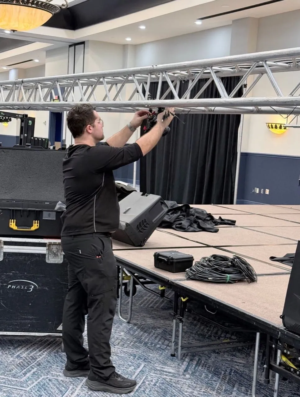 Phase 3 Live technician attaching fixtures to a lighting truss during stage setup.